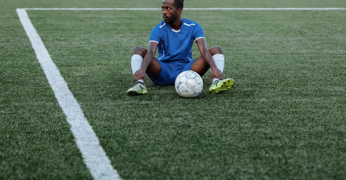 A soccer player in a blue uniform sits on a green field with a ball, reflecting focus and sportsmanship.