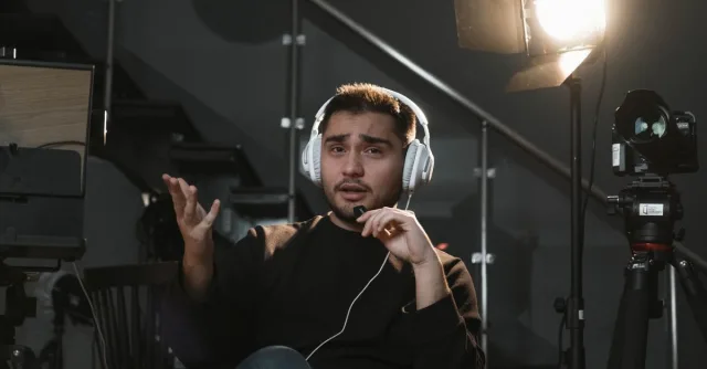Portrait of a man wearing headphones, sitting in a studio, gesturing towards the camera, with a microphone and lighting setup.
