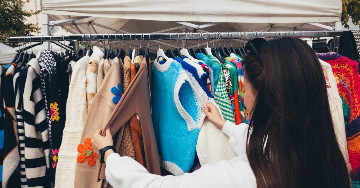 A woman browsing colorful vintage sweaters at an outdoor market stall.