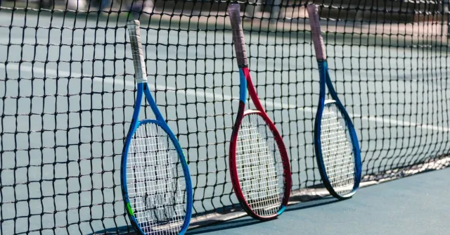 Three colorful tennis rackets leaning against a net on an outdoor tennis court.