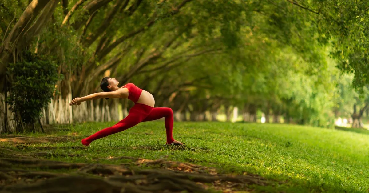 Pregnant woman in red practicing yoga outdoors in a lush green park, striking a pose.