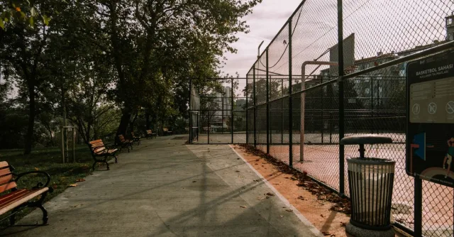 Empty outdoor basketball court with benches, trash bin, and chain-link fence on a sunny day.