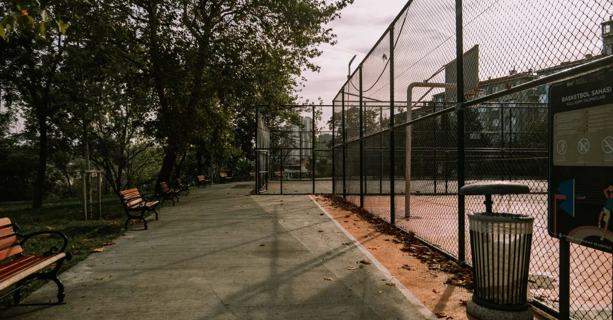 Empty outdoor basketball court with benches, trash bin, and chain-link fence on a sunny day.