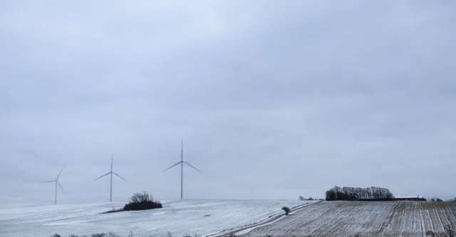 A serene snowy landscape in Randers, Denmark, featuring wind turbines under a cloudy sky.
