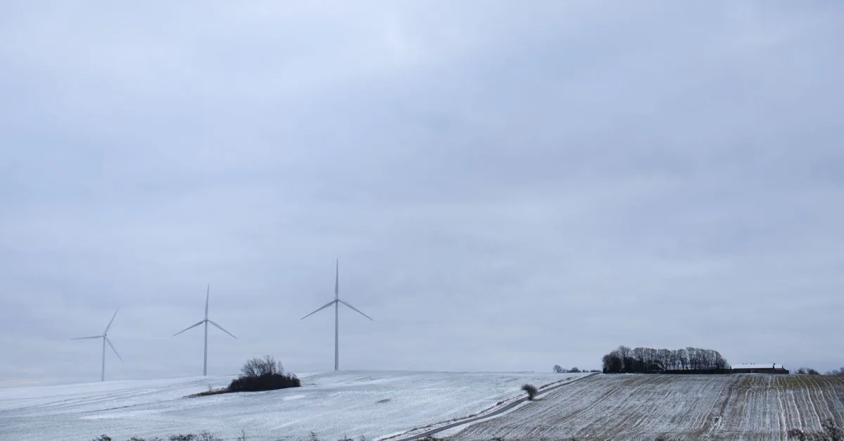 A serene snowy landscape in Randers, Denmark, featuring wind turbines under a cloudy sky.