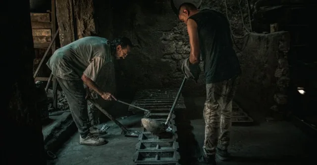 Two men working with molten metal in a dimly lit foundry setting.