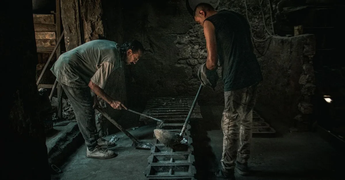 Two men working with molten metal in a dimly lit foundry setting.