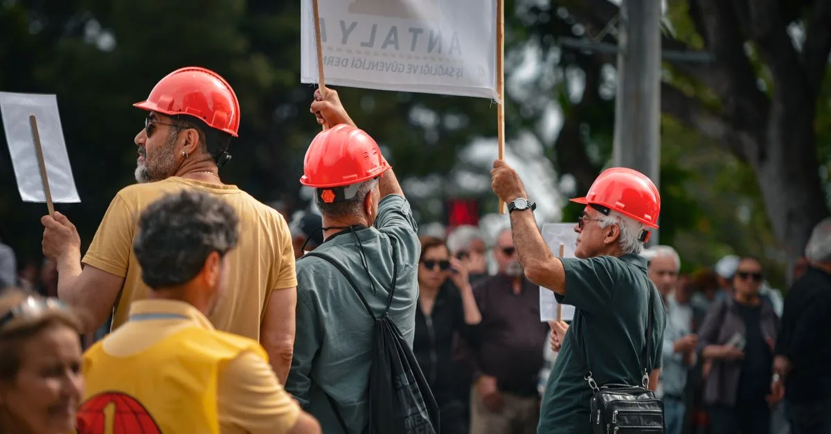 Protesters wearing red hard hats and holding signs gather in Antalya, Türkiye, highlighting activism.