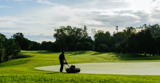 Groundskeeper mowing the green on a golf course during a sunny day, showcasing lush landscapes and maintenance.