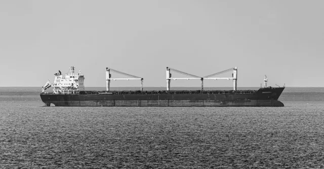 Black and white photo of a bulk carrier traversing calm open sea under a clear sky.