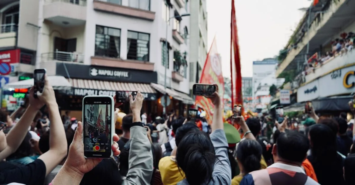 A vibrant street parade captured by many people using smartphones in a bustling urban scene.