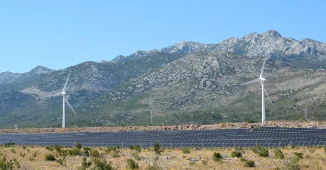 A scenic view of windmills and solar panels against mountainous terrain under a clear blue sky.