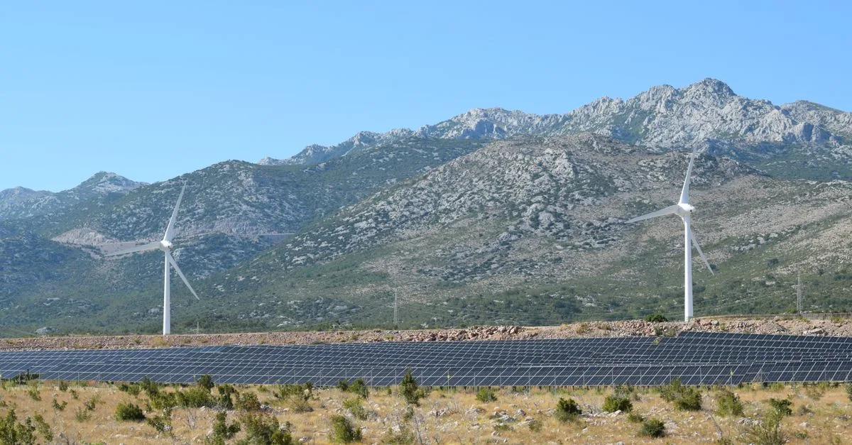 A scenic view of windmills and solar panels against mountainous terrain under a clear blue sky.