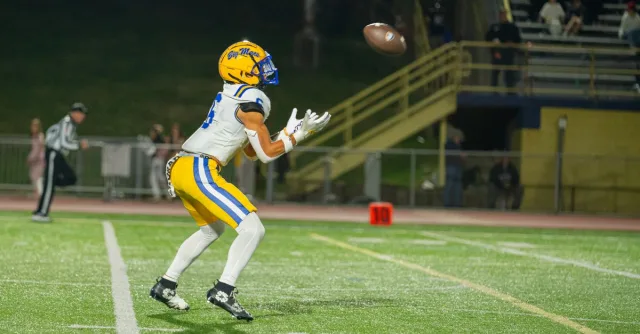 A high school football player wearing full gear catches a pass during an evening game on the field.