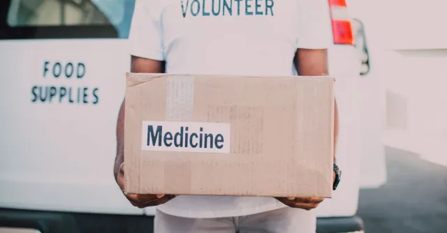 A volunteer holding a box labeled medicine near a delivery van marked food supplies.