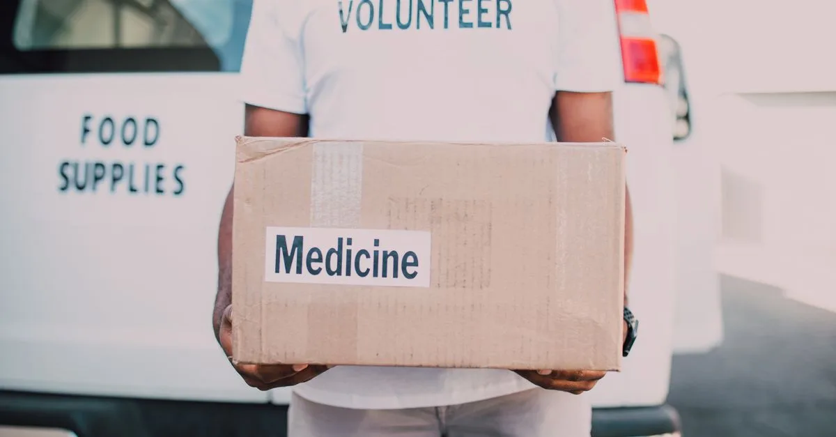 A volunteer holding a box labeled medicine near a delivery van marked food supplies.