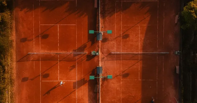Aerial shot of clay tennis courts with players during golden hour, casting long shadows.