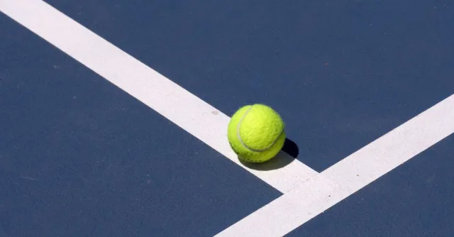 Close-up of a tennis ball on a blue court with white lines under sunlight.