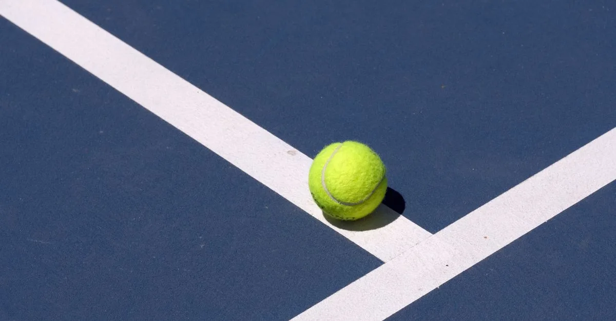 Close-up of a tennis ball on a blue court with white lines under sunlight.