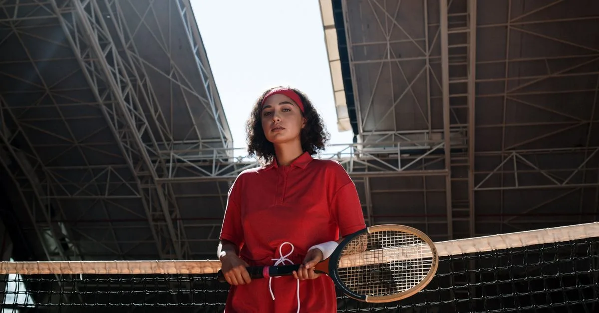A woman in a red outfit stands with a tennis racket indoors, exuding vintage fashion vibes.
