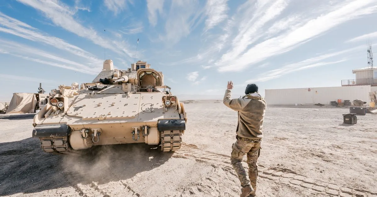 Bradley Fighting Vehicle and soldier in desert training operation under clear sky.