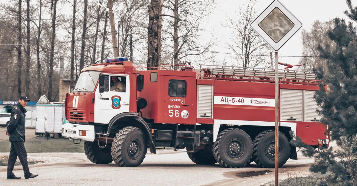 A firetruck is parked on a road with a man in uniform walking nearby, trees in the background.
