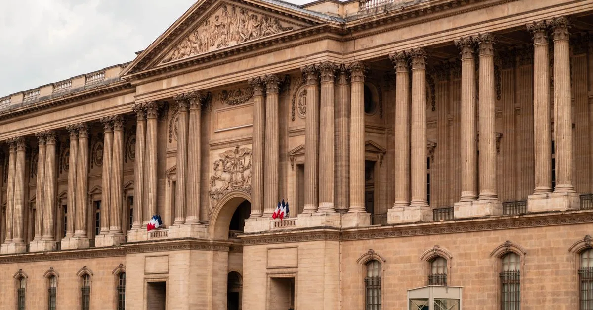 Front view of the iconic Louvre Museum facade, Paris, with French flags displayed.