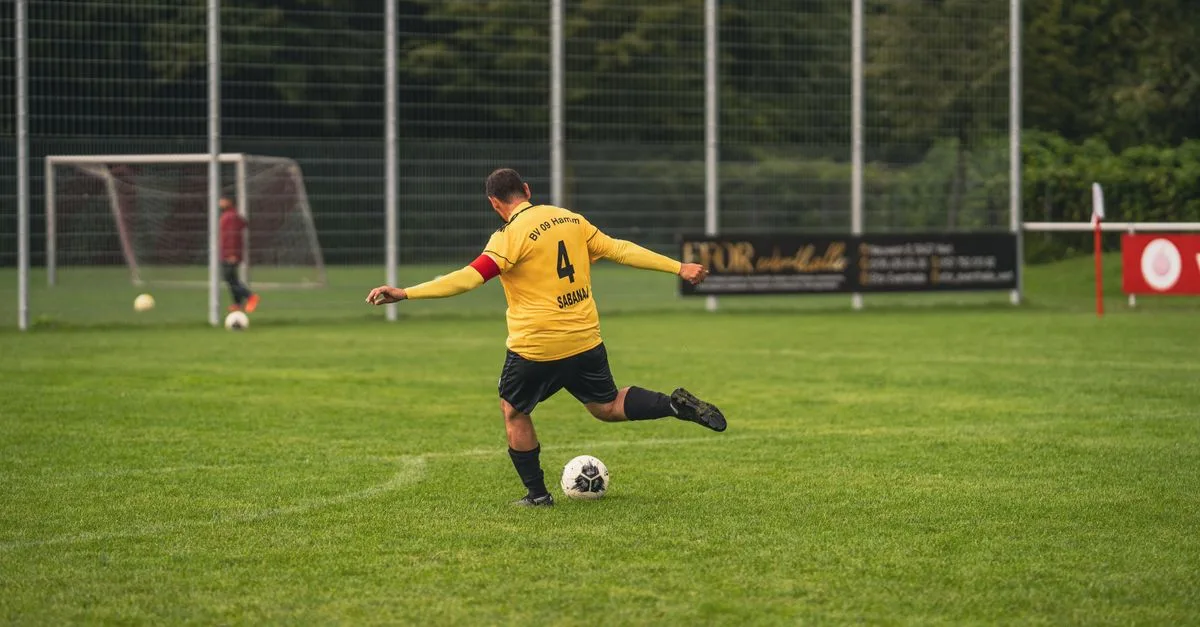 An amateur soccer player in yellow jersey kicks a ball on a grassy field during the day.