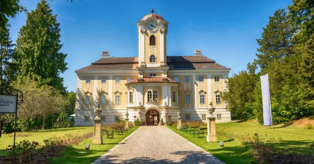 A grand European building with a clock tower set amidst lush greenery under a clear sky.