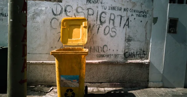 Yellow recycle bin with graffiti on wall in Pacasmayo, Perú, highlighting urban decay and sustainability.