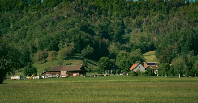 Idyllic countryside scene with farmhouses, green pastures, and lush forested hills under a clear blue sky.