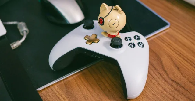 A charming cat figurine sits atop a gaming controller, beside a computer mouse, on a wooden desk.