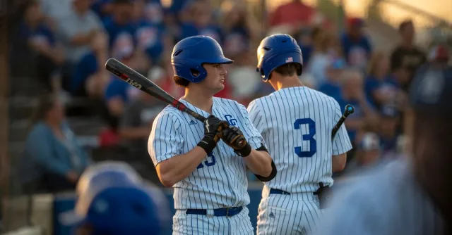 Two baseball players in uniform, standing with bats during a game at sunset.
