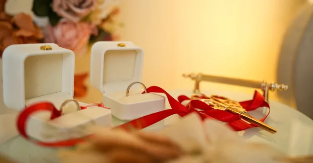 Close-up of wedding rings in boxes with decorative flowers and ribbons on a glass table.