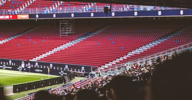 Panoramic view of stadium with empty red seats and crowd gathered for a live match.