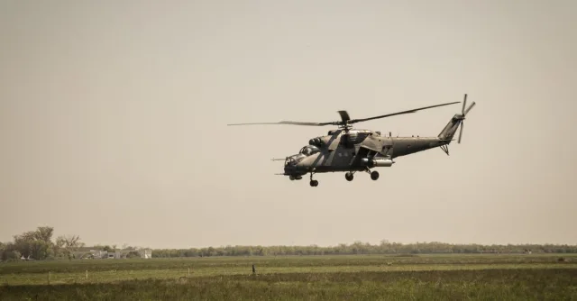 A military helicopter flies over a vast green grassland under a clear sky.