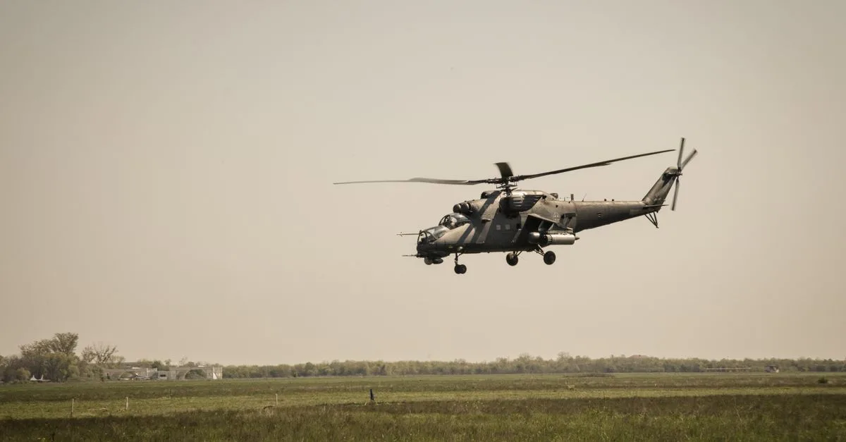 A military helicopter flies over a vast green grassland under a clear sky.