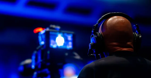 A videographer wearing headphones captures an event with professional video equipment in a dimly lit setting.