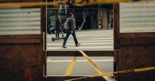 Men crossing a city street with caution tape in the foreground, including a person on an electric scooter.