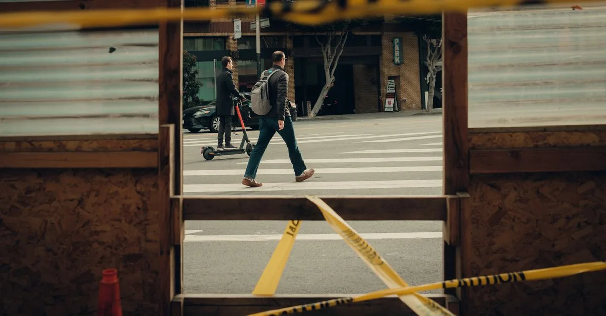 Men crossing a city street with caution tape in the foreground, including a person on an electric scooter.