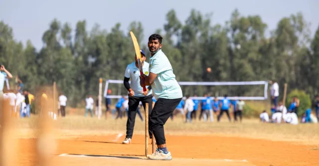 A cricketer prepares to bat during an outdoor game on a sunny day, with spectators in the background.