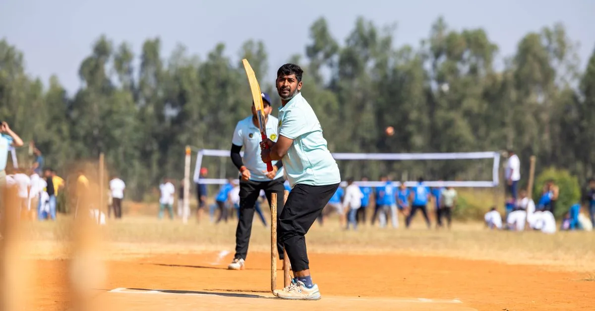 A cricketer prepares to bat during an outdoor game on a sunny day, with spectators in the background.
