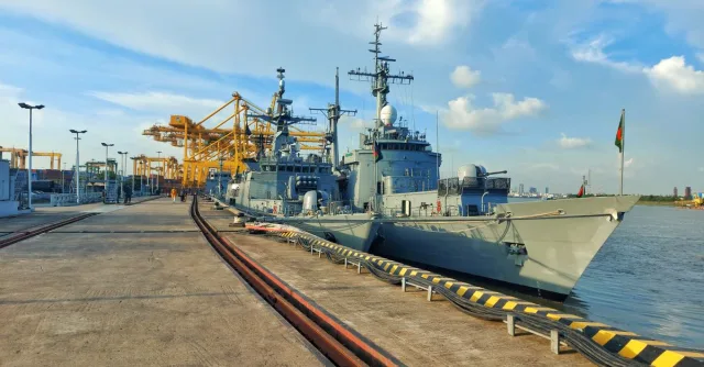 Navy vessels docked at a bustling city port with cranes and clear blue skies.
