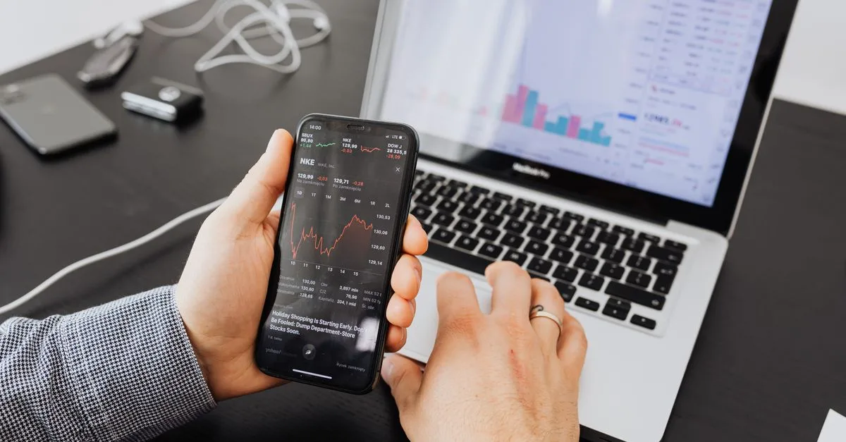 Person analyzes stock data with a smartphone and laptop, indoors.