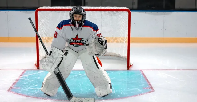 Focused ice hockey goalkeeper stands ready in front of the goal, equipped with helmet and pads on an indoor ice rink.