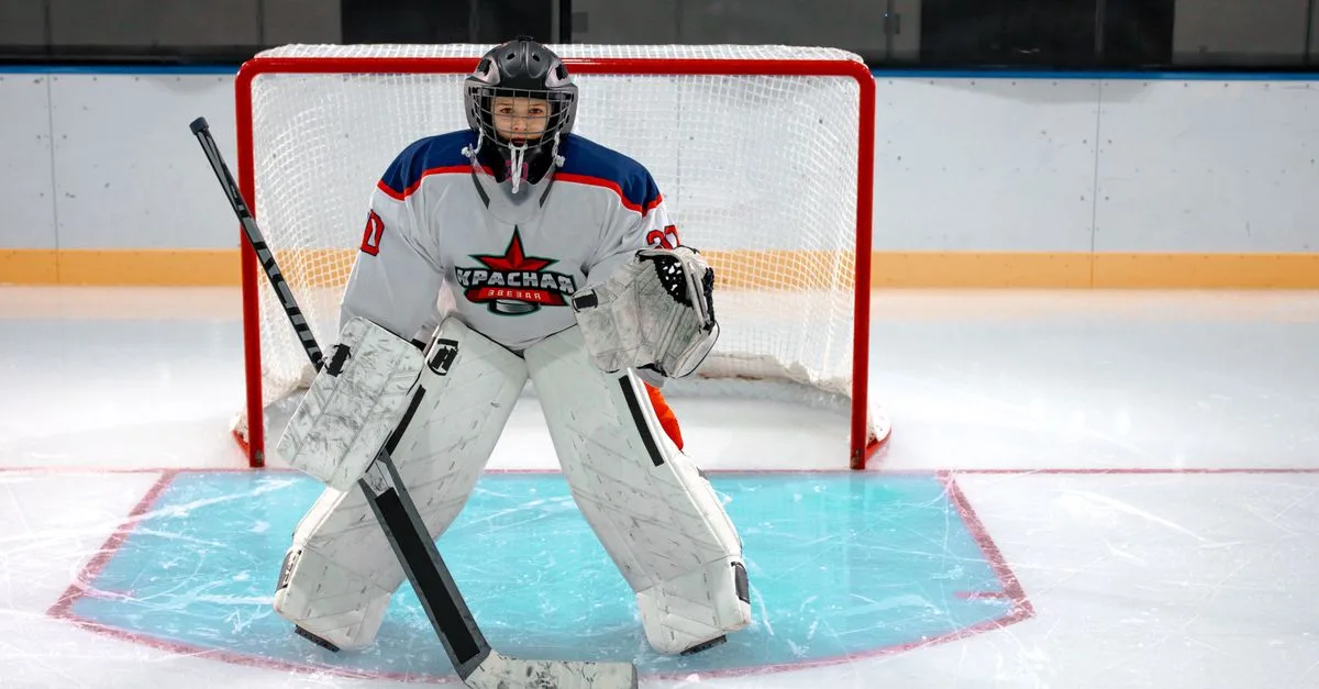 Focused ice hockey goalkeeper stands ready in front of the goal, equipped with helmet and pads on an indoor ice rink.