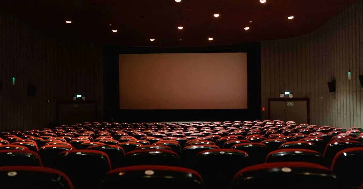 A dimly lit empty cinema theater with rows of red chairs facing a projector screen.