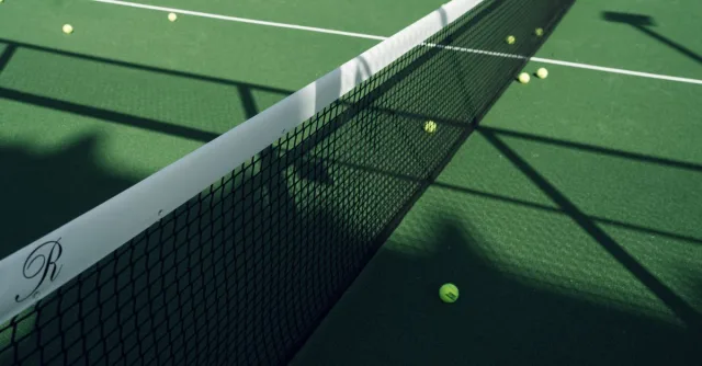 Tennis court net casting shadows on a green surface with scattered tennis balls.