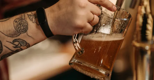Close-up of a tattooed hand pouring beer into a glass mug from a draft tap.