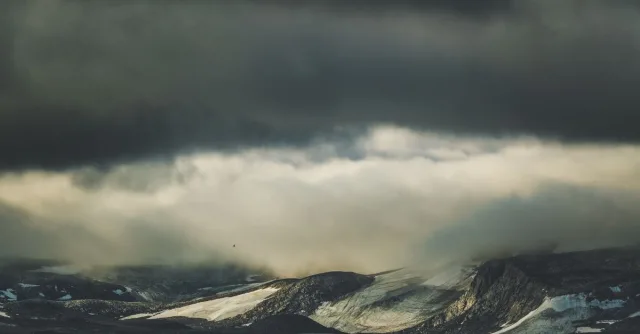A moody landscape of mountains beneath a dramatic overcast sky full of thick clouds.